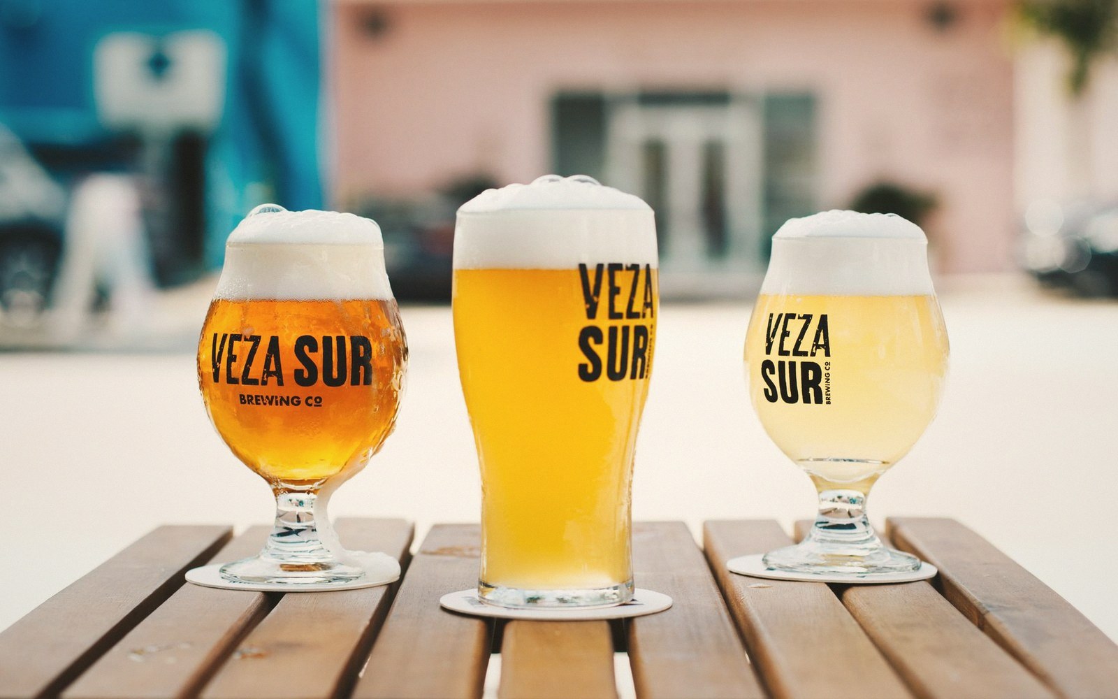 Three foam-topped Veza Sur beers in branded glasses lined up on an outdoor table