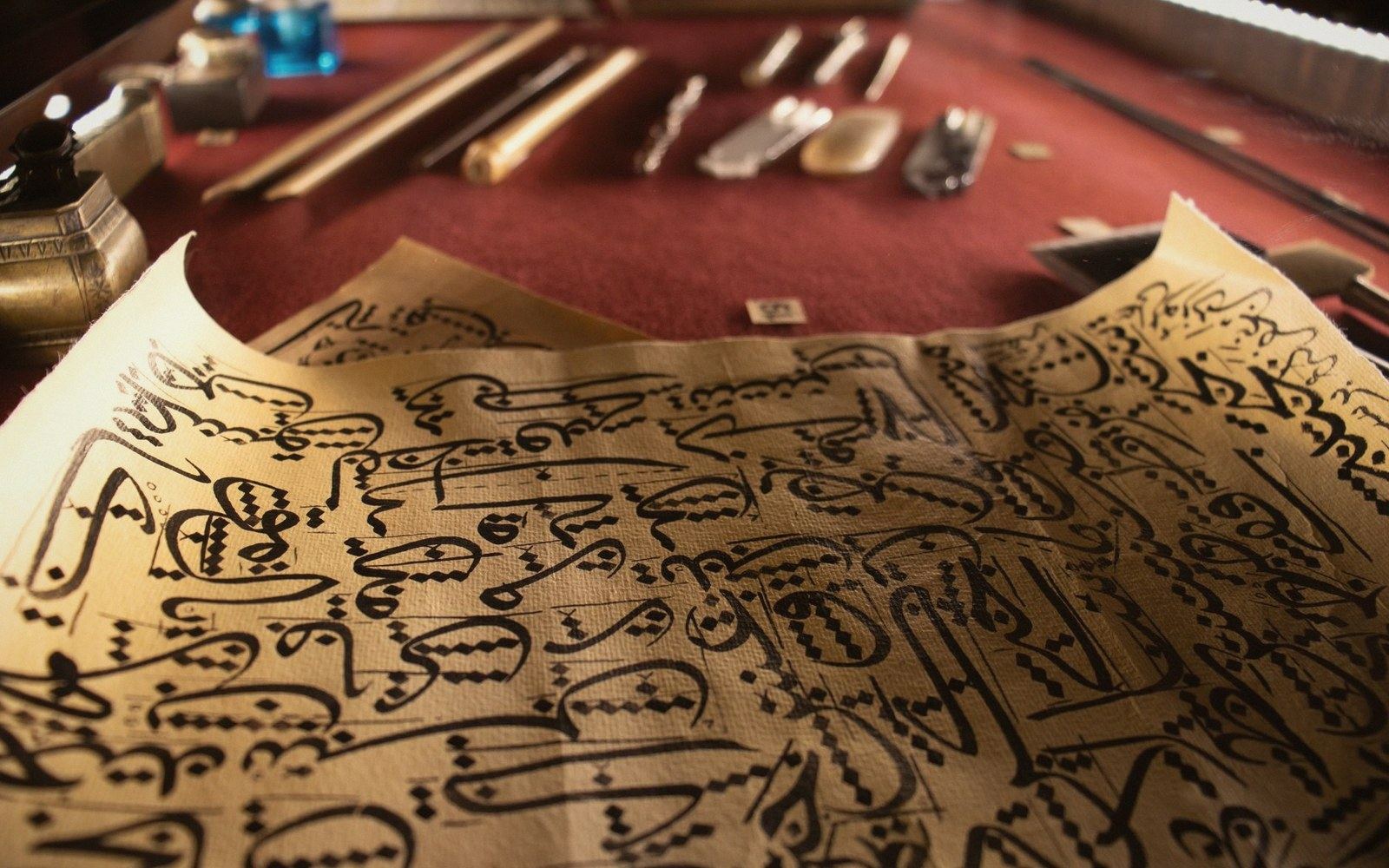 Arabic calligraphy practice sheet lies beside reed pens and ink bottles on a desk